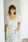 A woman smiling and wearing a white smocked dress with a muted green floral pattern, standing in a brightly lit indoor setting.