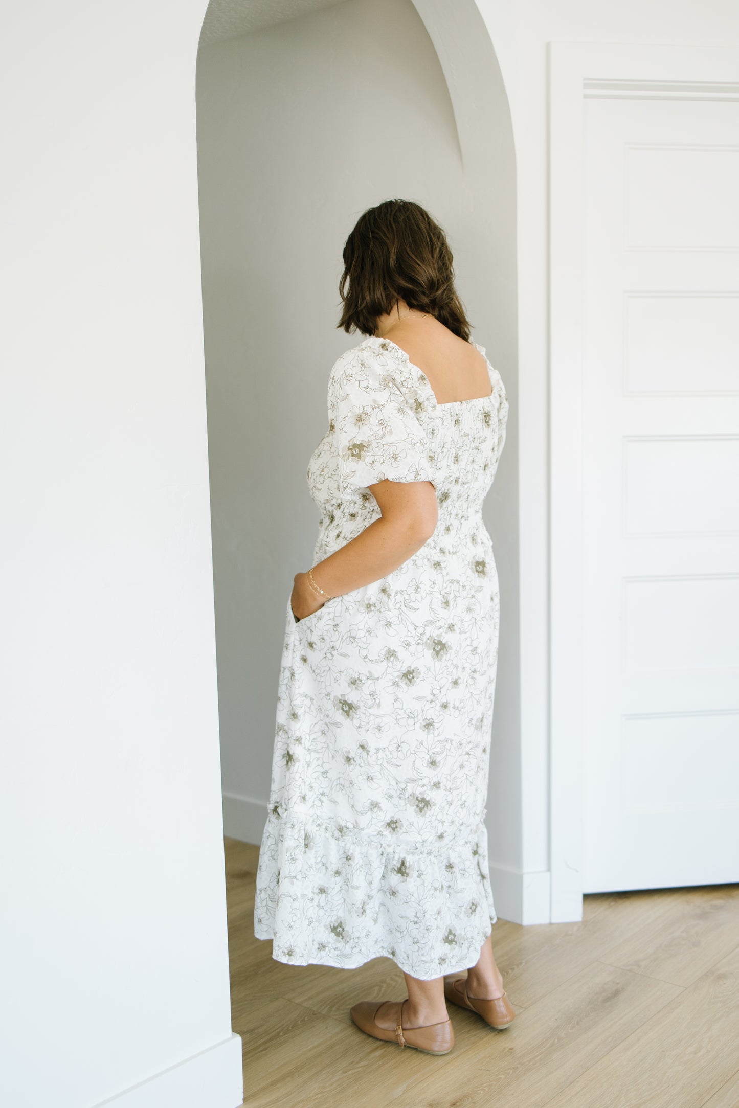 A woman wearing a white floral midi dress with puff sleeves and side pockets, standing in a bright white room with light wood floors.