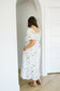 A woman wearing a white floral midi dress with puff sleeves and side pockets, standing in a bright white room with light wood floors.