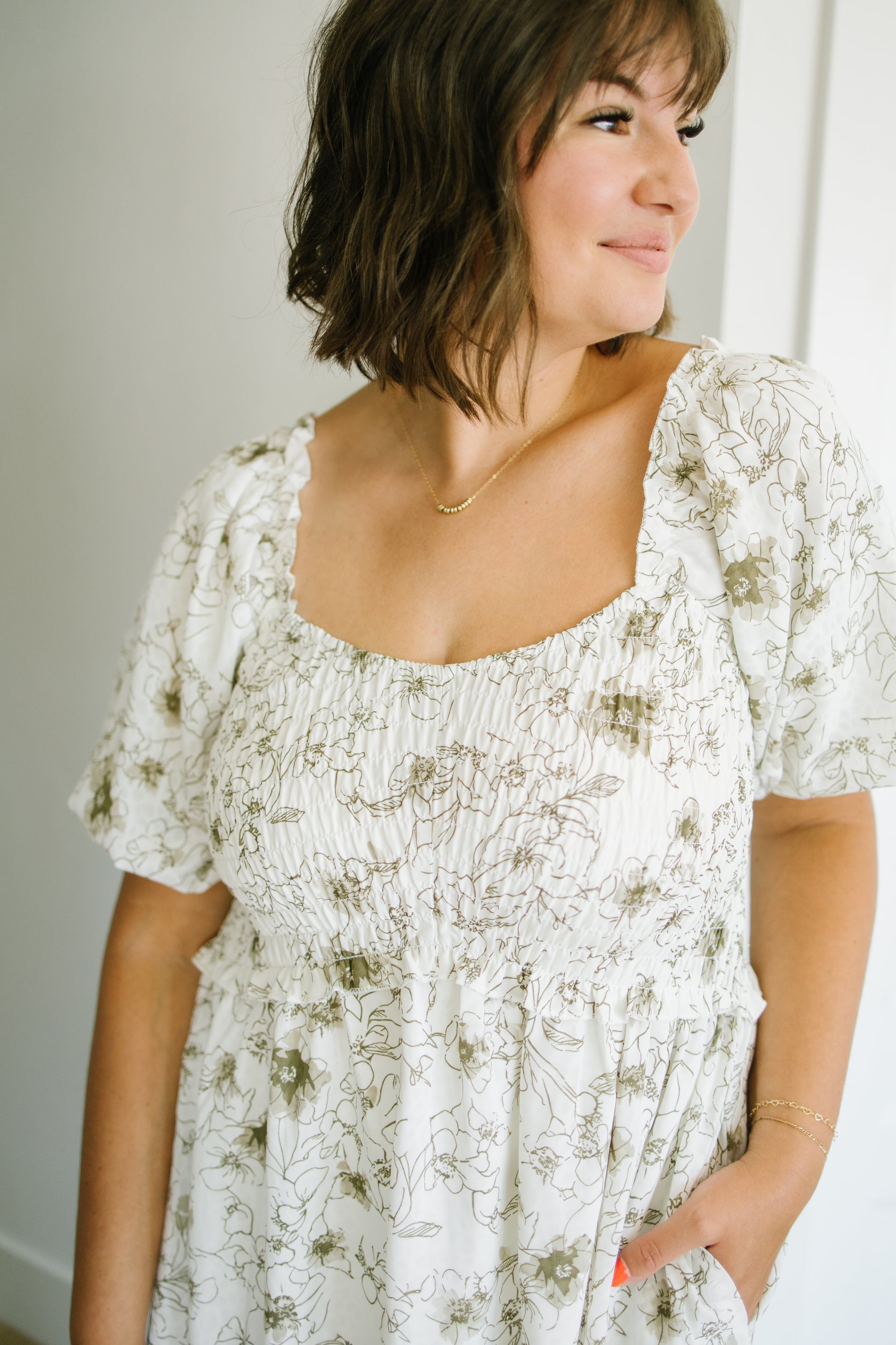 A woman wearing a white smocked dress with a muted green floral line print, featuring puff sleeves and a square neckline.