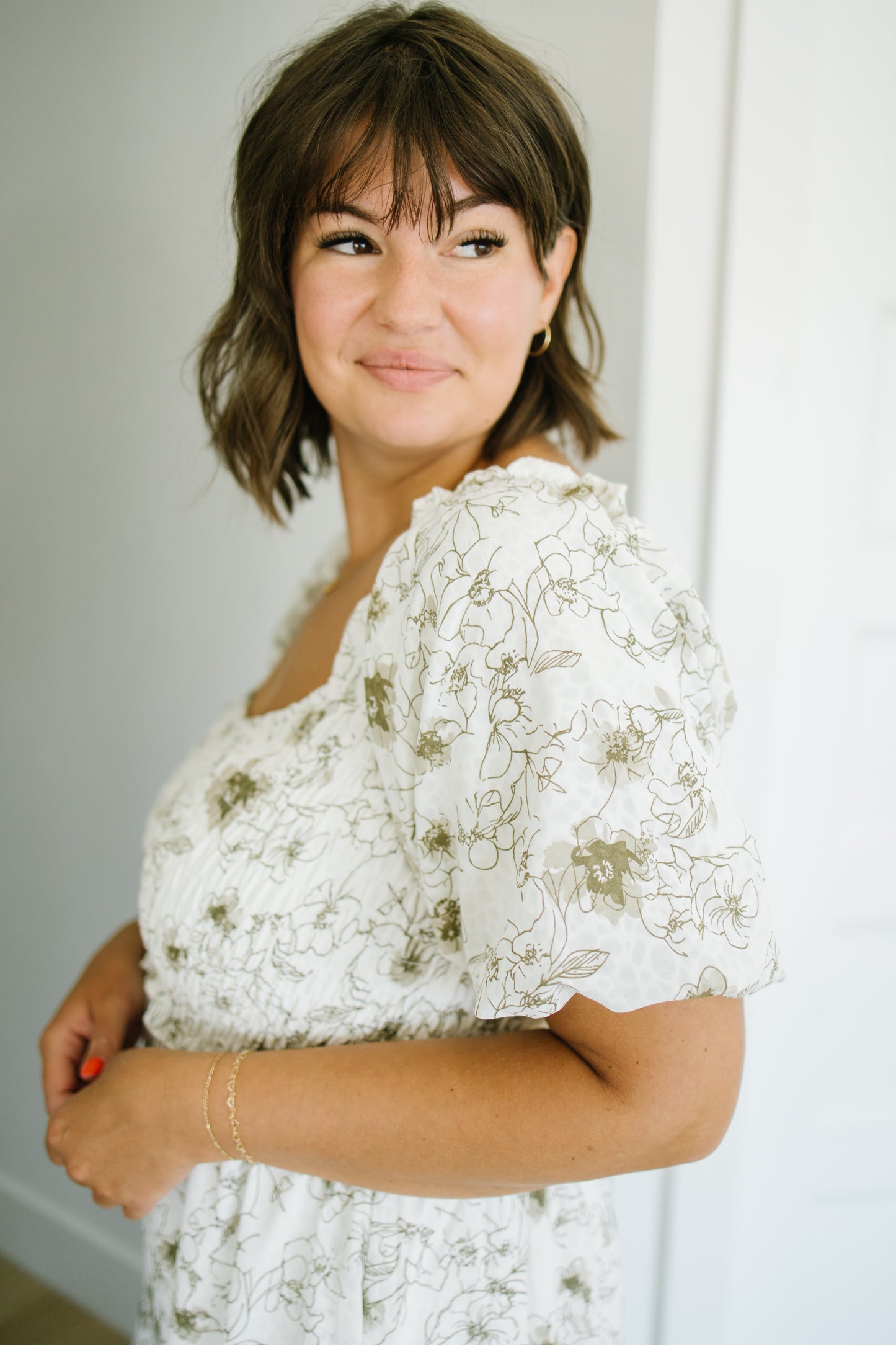 A woman with short brown hair and bangs wearing a white puff-sleeve dress with a delicate olive green floral pattern, posing in a brightly lit indoor setting.