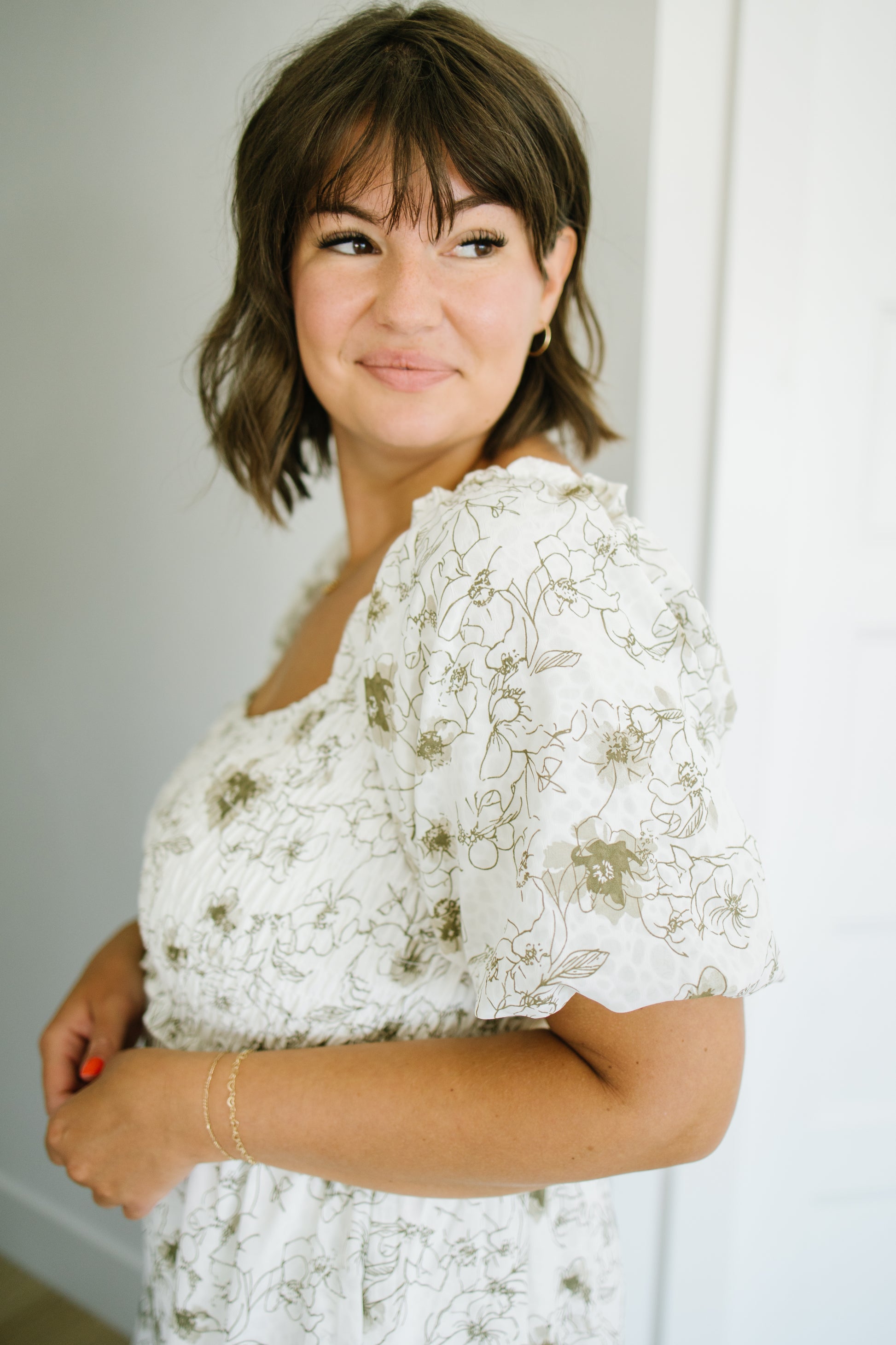 A woman with short brown hair and bangs wearing a white puff-sleeve dress with a delicate olive green floral pattern, posing in a brightly lit indoor setting.