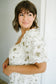 A woman with short brown hair and bangs wearing a white puff-sleeve dress with a delicate olive green floral pattern, posing in a brightly lit indoor setting.