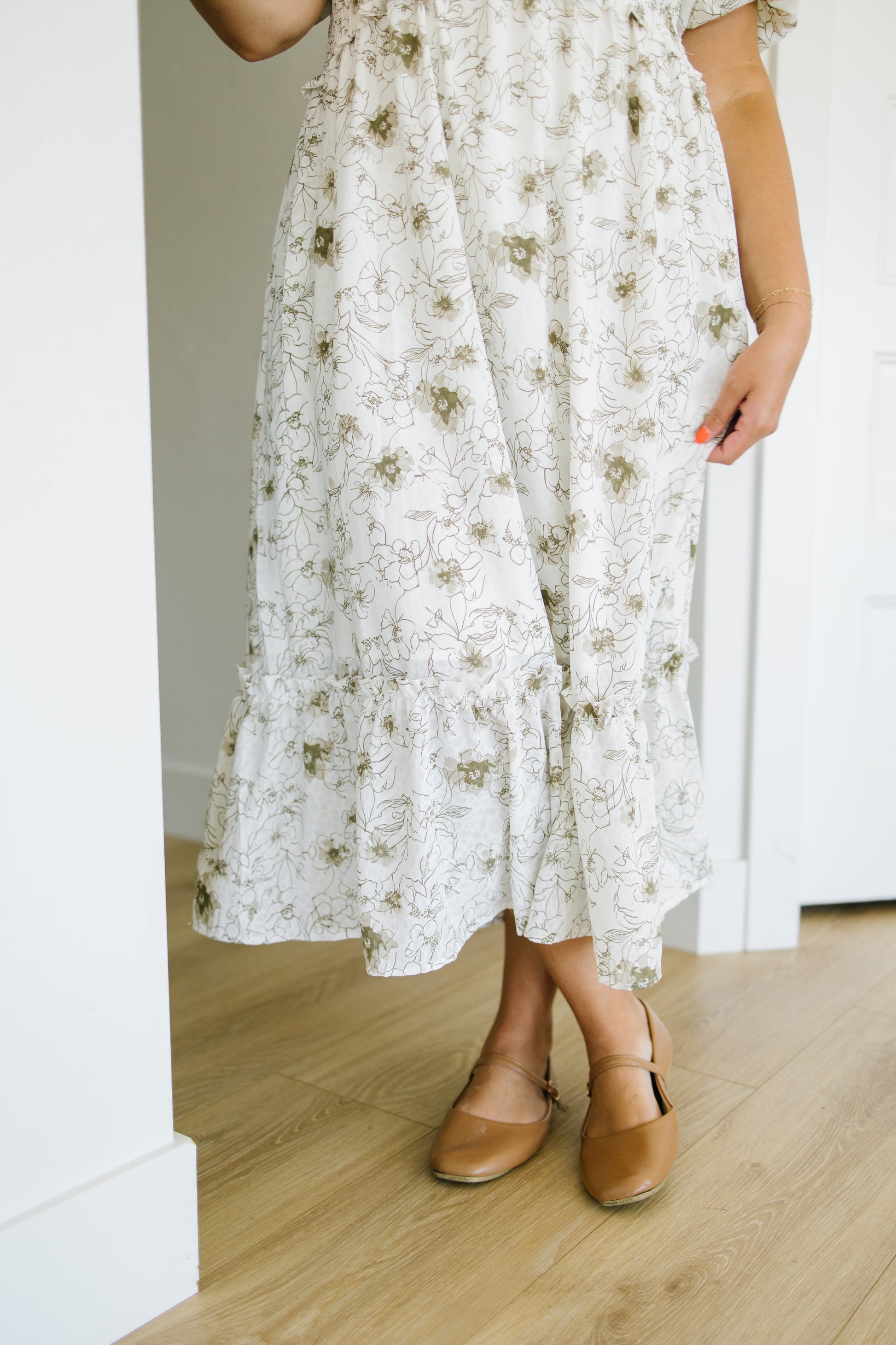 A person wearing a white midi dress with a muted olive green floral pattern, paired with tan flat shoes, standing in a brightly lit room with light wood floors and white walls.
