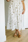 A person wearing a white midi dress with a muted olive green floral pattern, paired with tan flat shoes, standing in a brightly lit room with light wood floors and white walls.