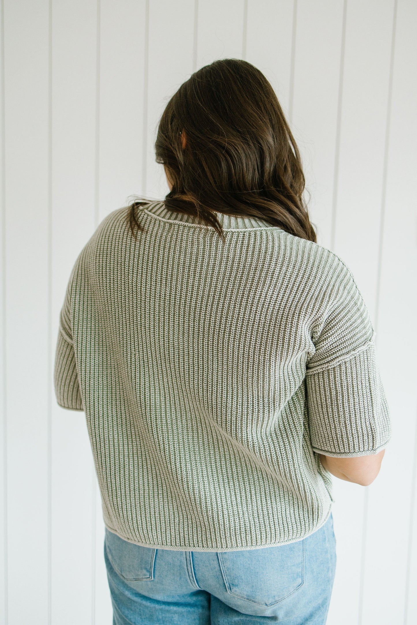Person wearing a light green ribbed sweater and blue jeans against a white paneled wall.