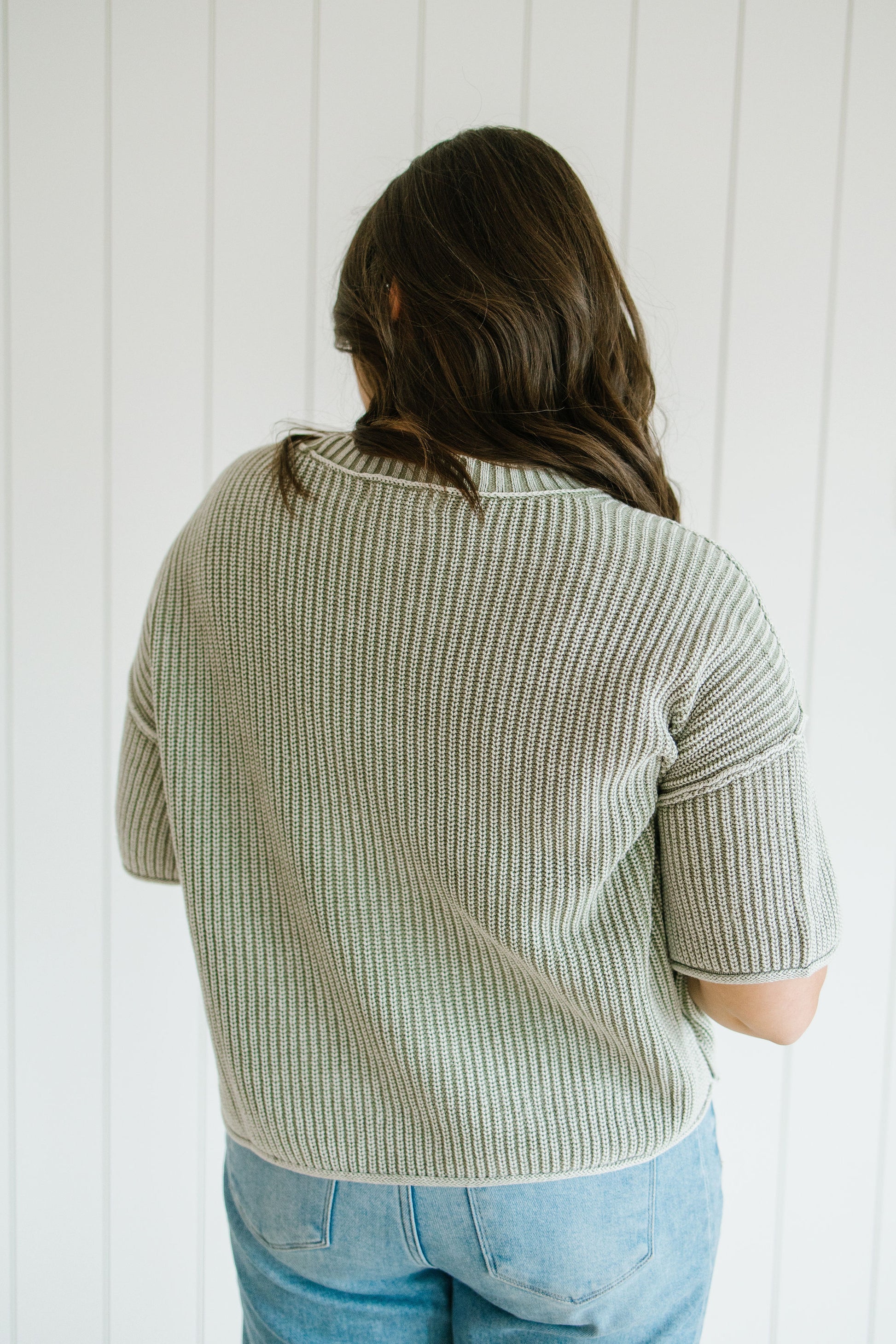 Person wearing a light green ribbed sweater and blue jeans against a white paneled wall.