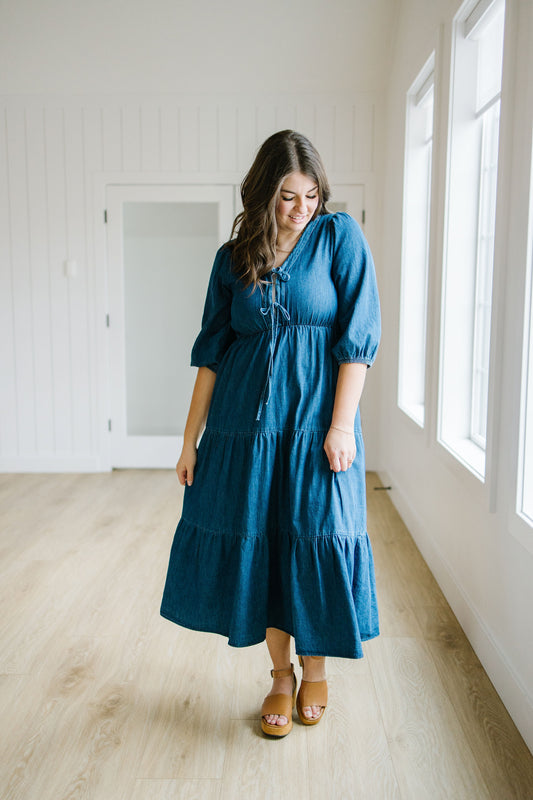 Woman wearing a blue denim tiered dress standing in a bright room with large windows.
