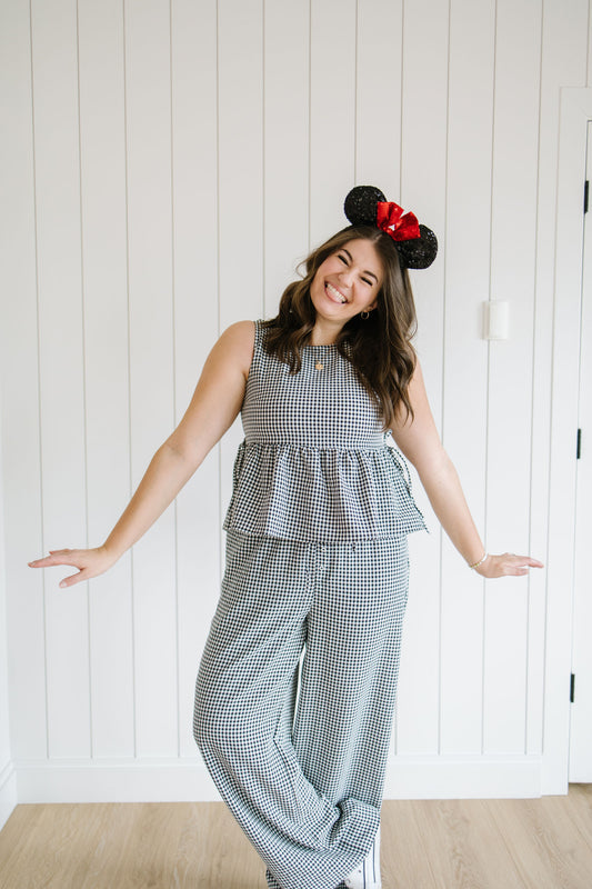 Woman wearing a black gingham checkered matching set with a mouse
headband against a white paneled wall.
