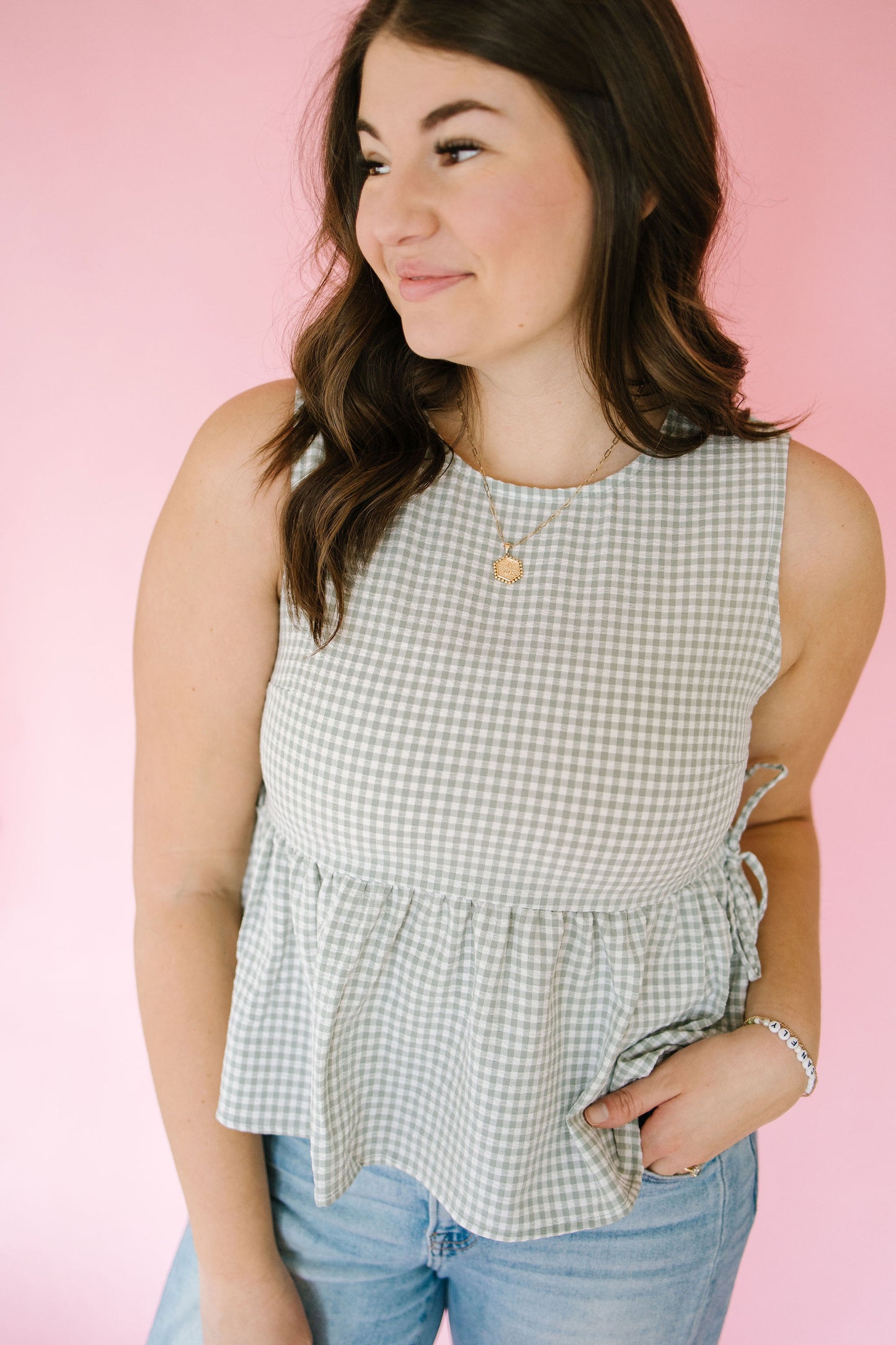 Woman wearing a sage green gingham  sleeveless top and jeans against a pink background
