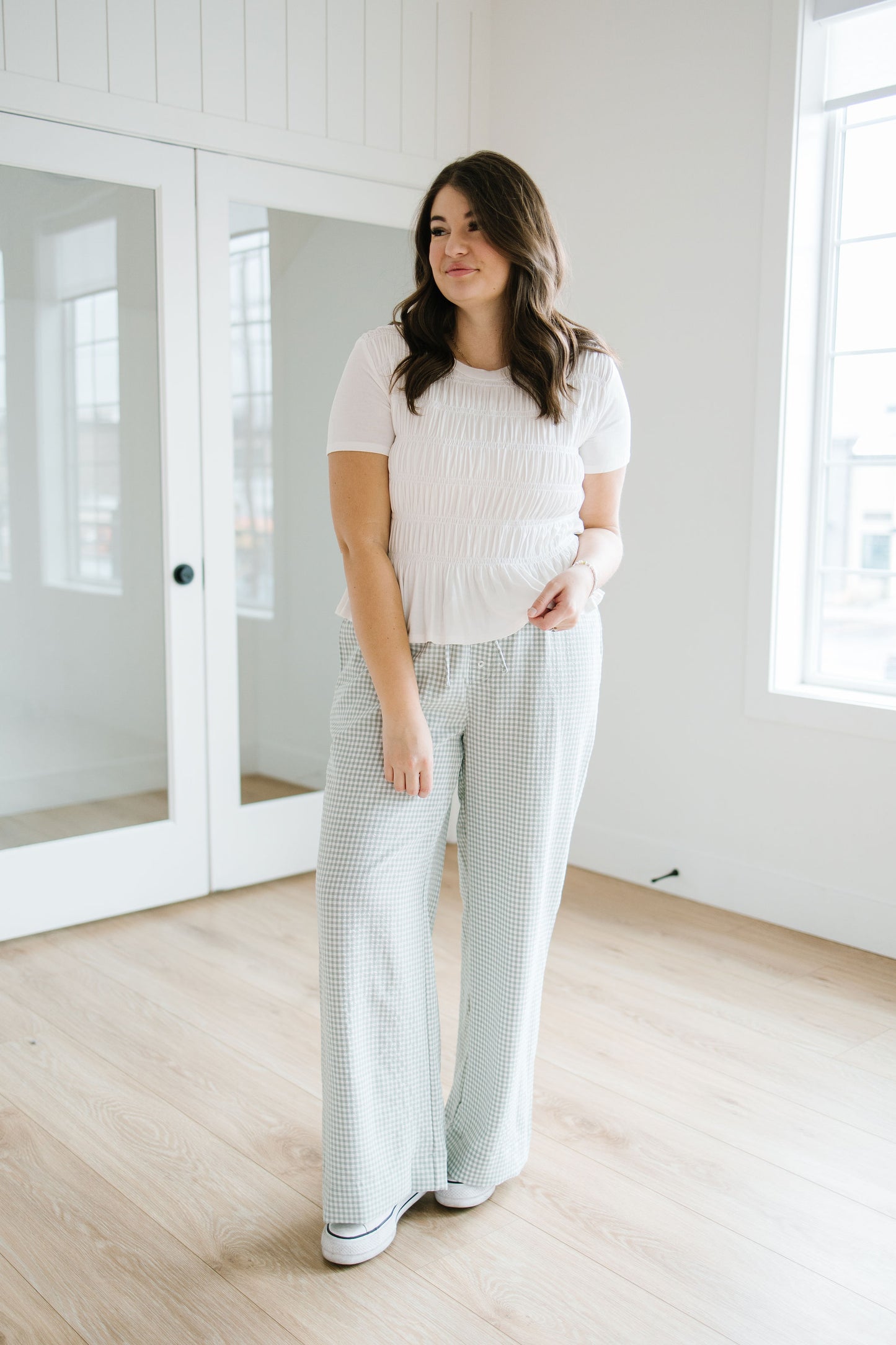Woman in a white top and light checkered pants standing in a minimalistic room with white walls and wooden floor.