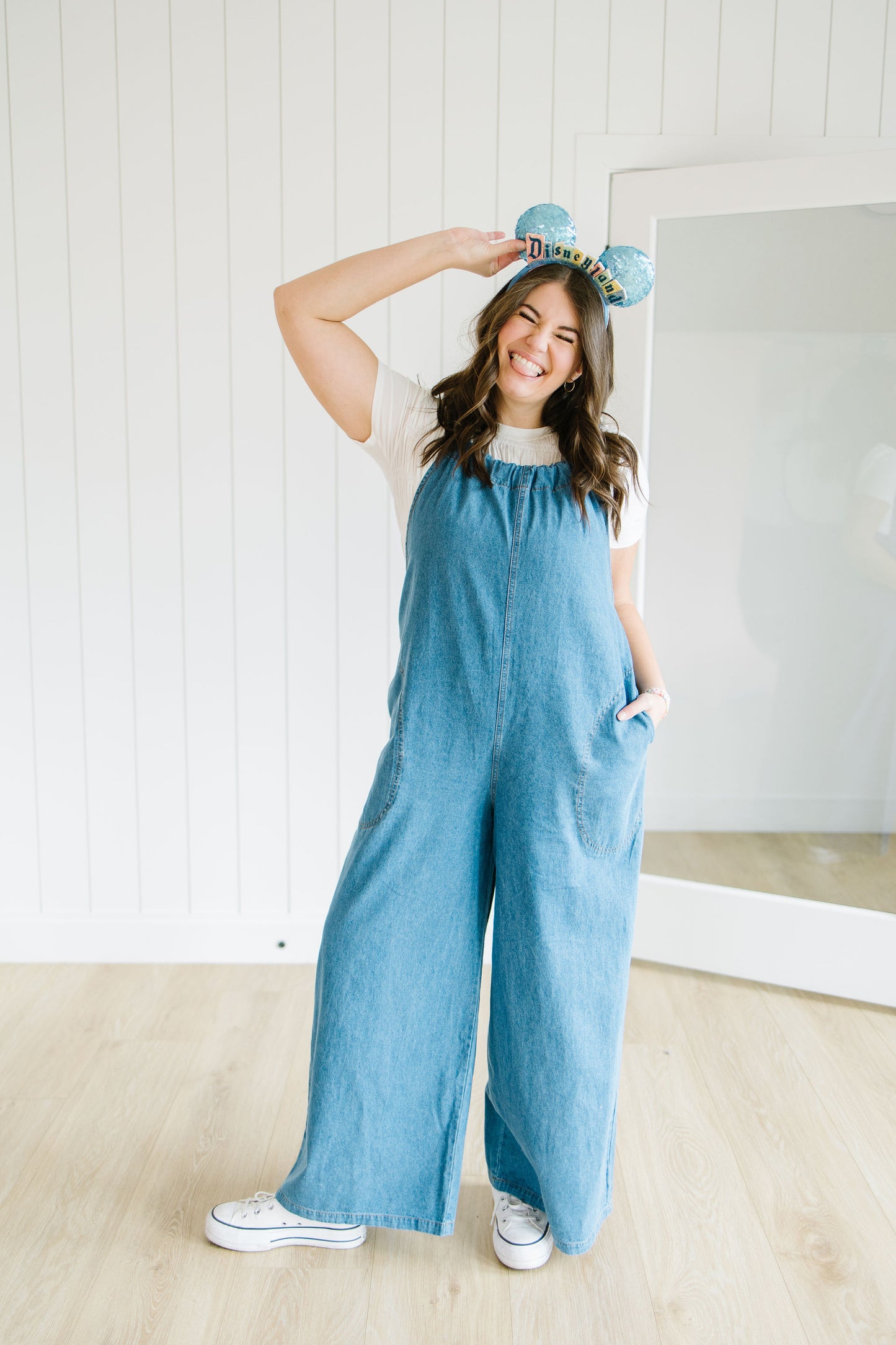 Woman wearing a blue denim wide leg jumpsuit standing in a room with a mirror.