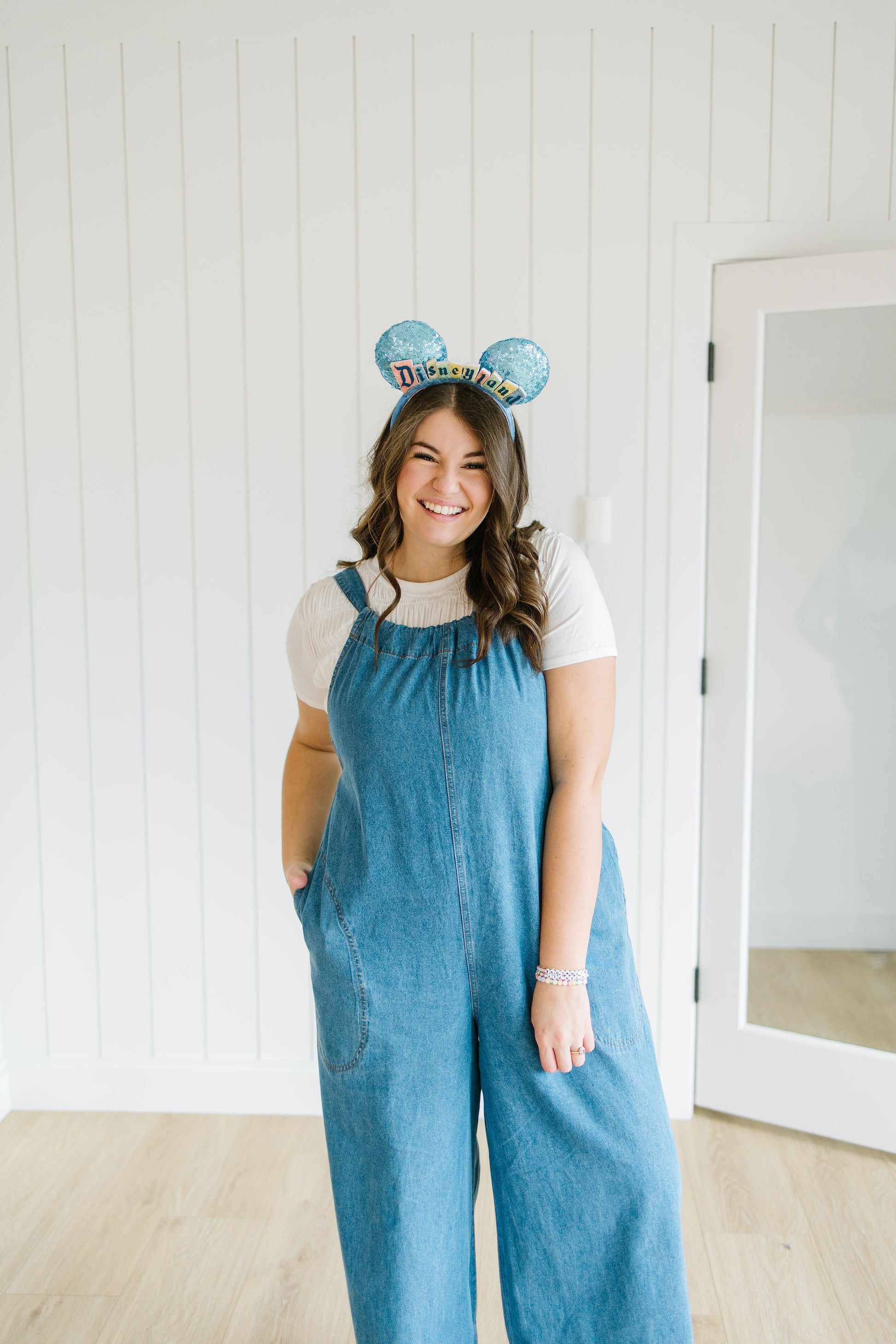 Person wearing blue overalls and a white t-shirt with a decorative Disneyland headband indoors.