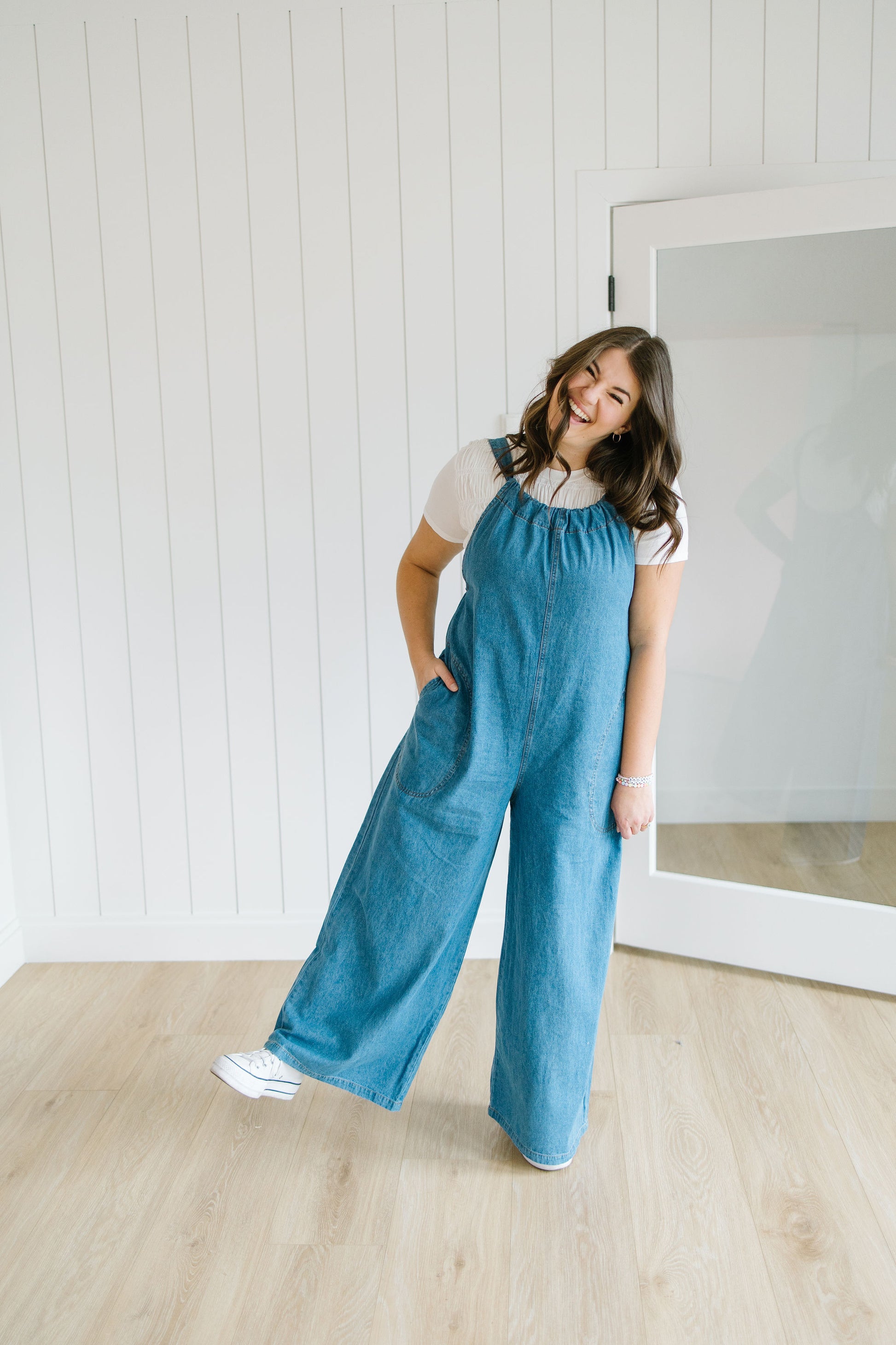 Woman wearing a blue jumpsuit standing in a room with a white wall and wooden floor.