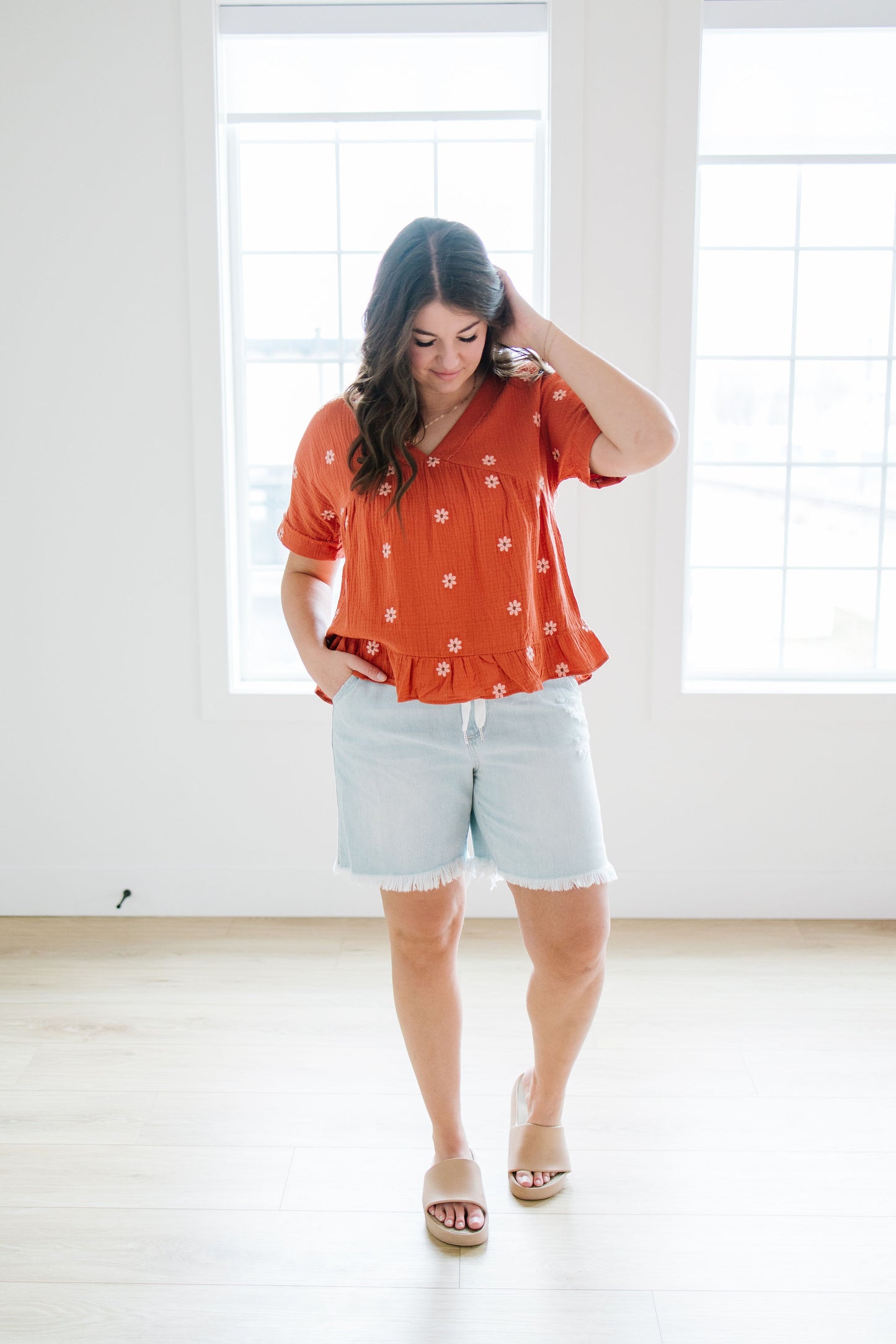 Woman wearing an orange blouse with white embroidered floral patterns and light blue shorts in a bright room.