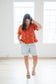 Woman wearing an orange blouse with white embroidered floral patterns and light blue shorts in a bright room.