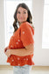 Woman wearing an orange blouse with white floral patterns in a bright room.