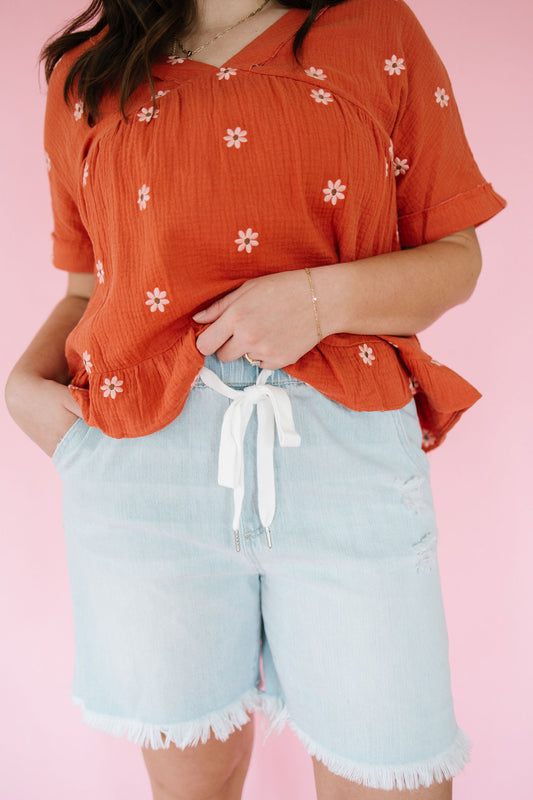 Person wearing an orange floral top and light blue shorts against a pink background