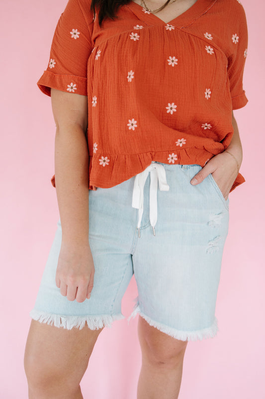 Person wearing an orange floral top and light blue shorts on a pink background