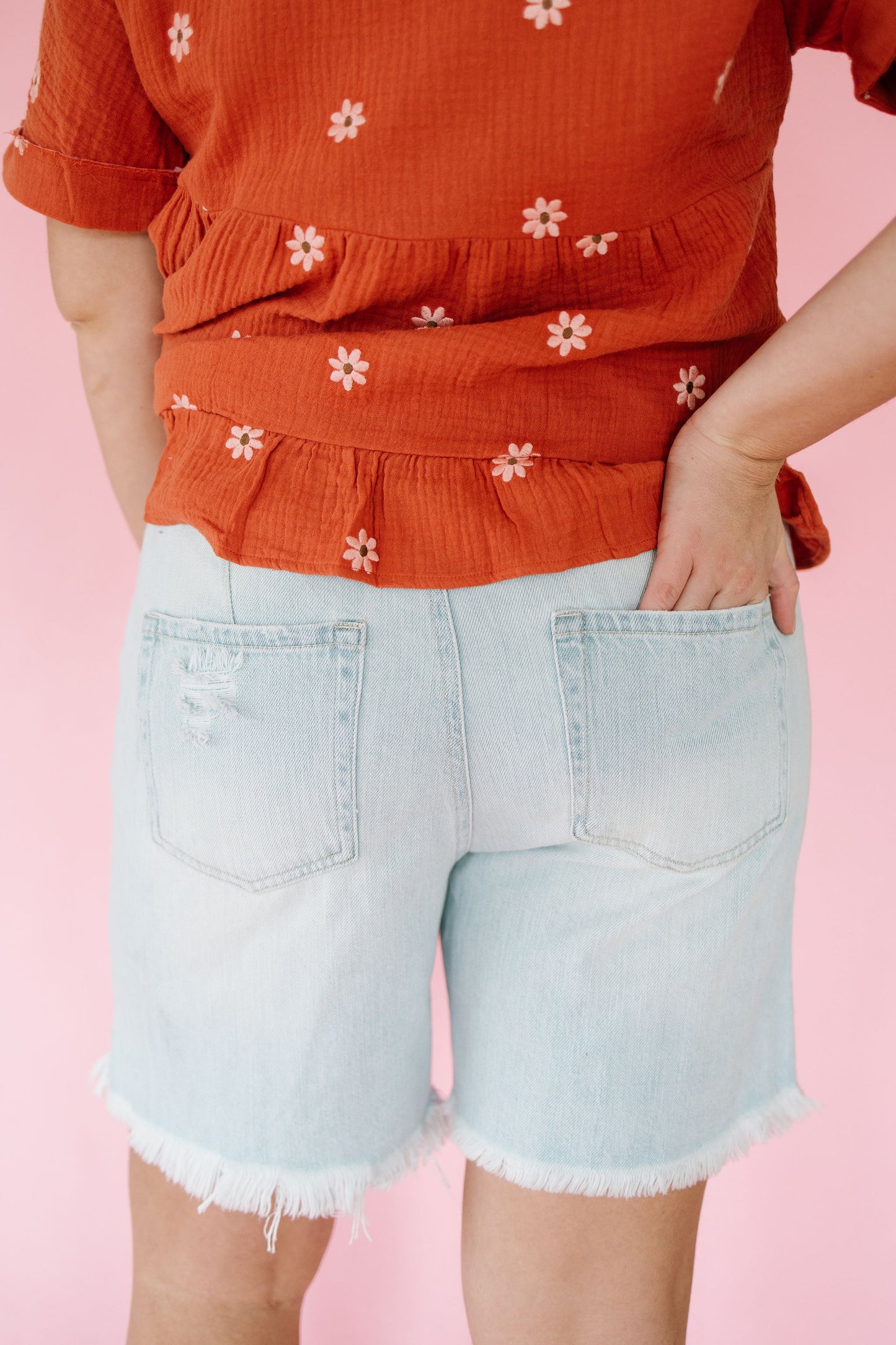 Person wearing an orange top with white floral patterns and light blue denim shorts on a pink background