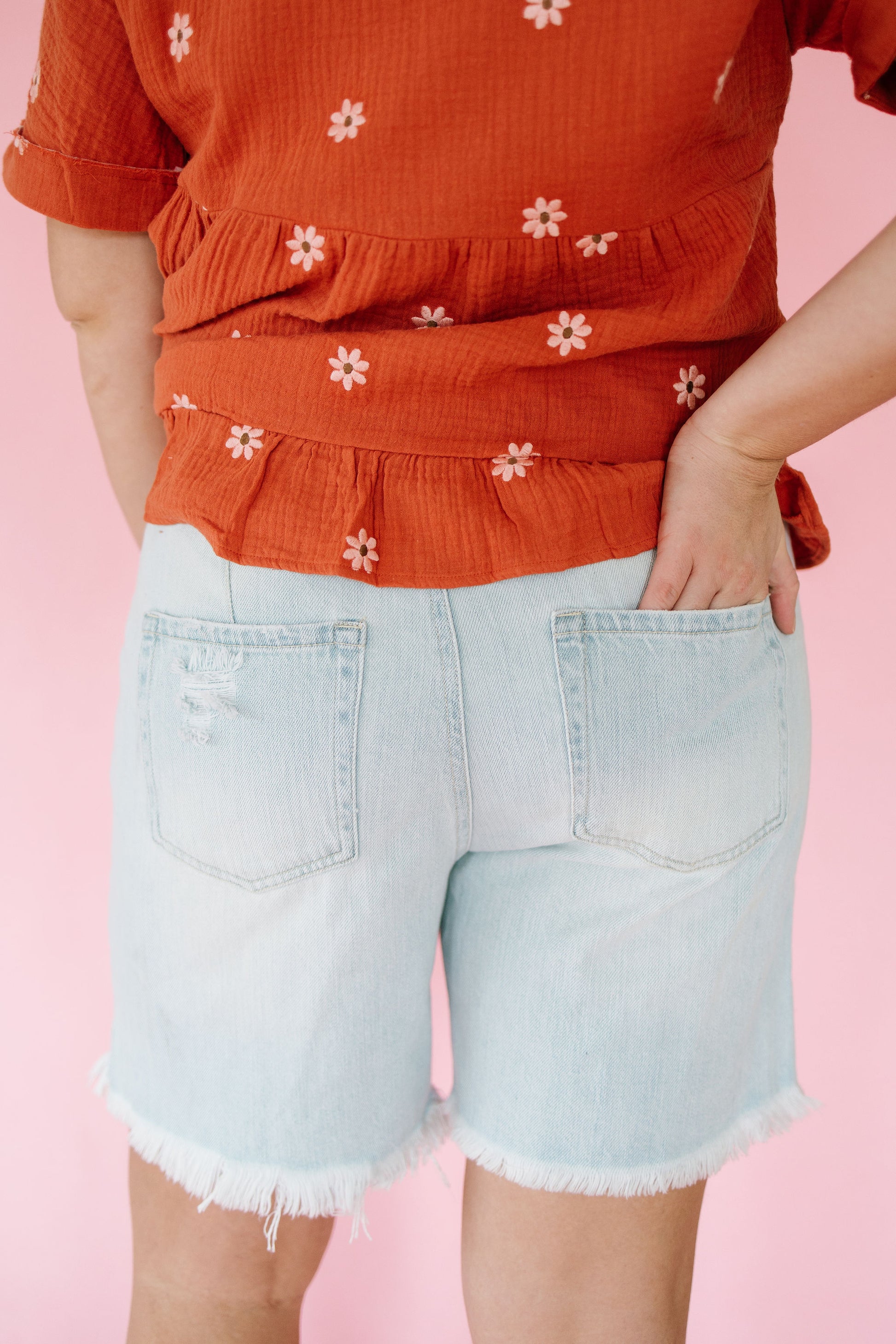 Person wearing an orange top with white floral patterns and light blue denim shorts on a pink background