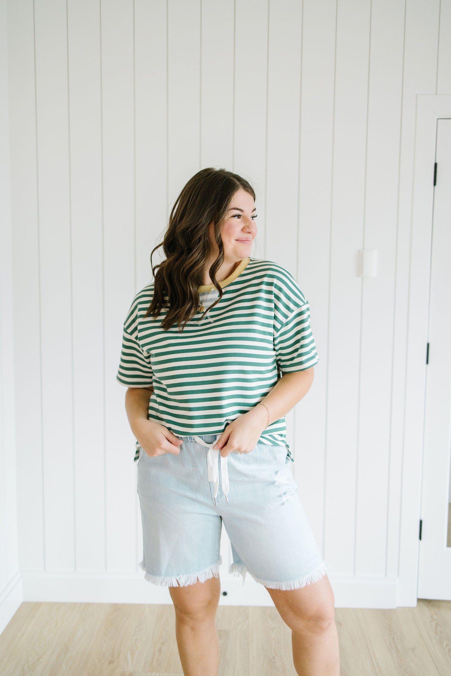 Woman wearing a green and white striped shirt and light blue shorts standing against a white paneled wall.