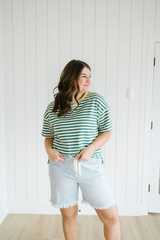 Woman wearing a green and white striped shirt and light blue shorts standing against a white paneled wall.
