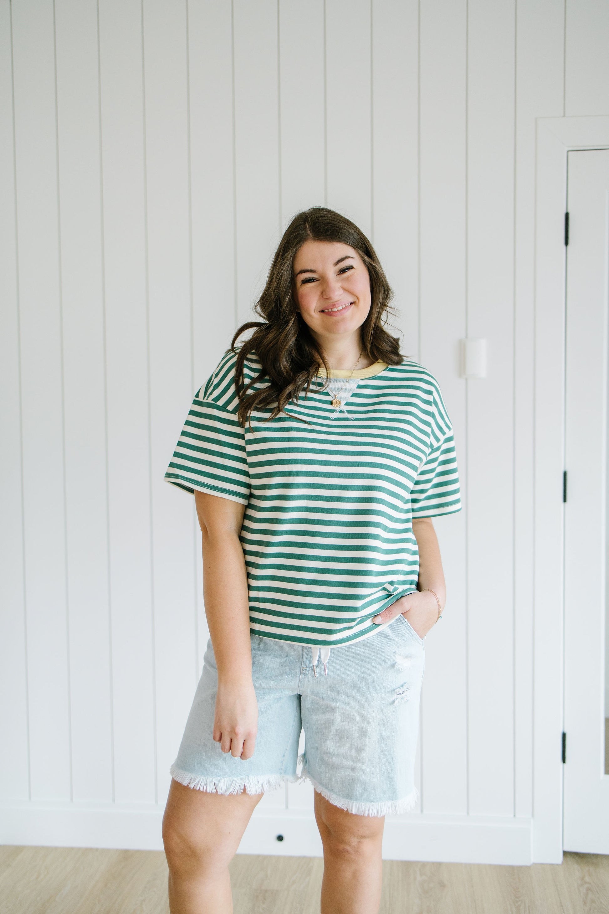 Woman wearing a green and white striped shirt and denim shorts standing against a white paneled wall.